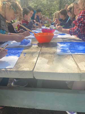 Students adding blue paint to paper while sitting at an outdoor picnic table